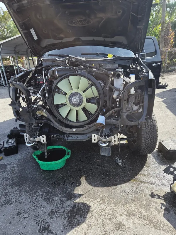 Front of a Ford pickup with the hood up and the front bumper and grille removed, exposing the engine and cooling fan, with a drain pan underneath