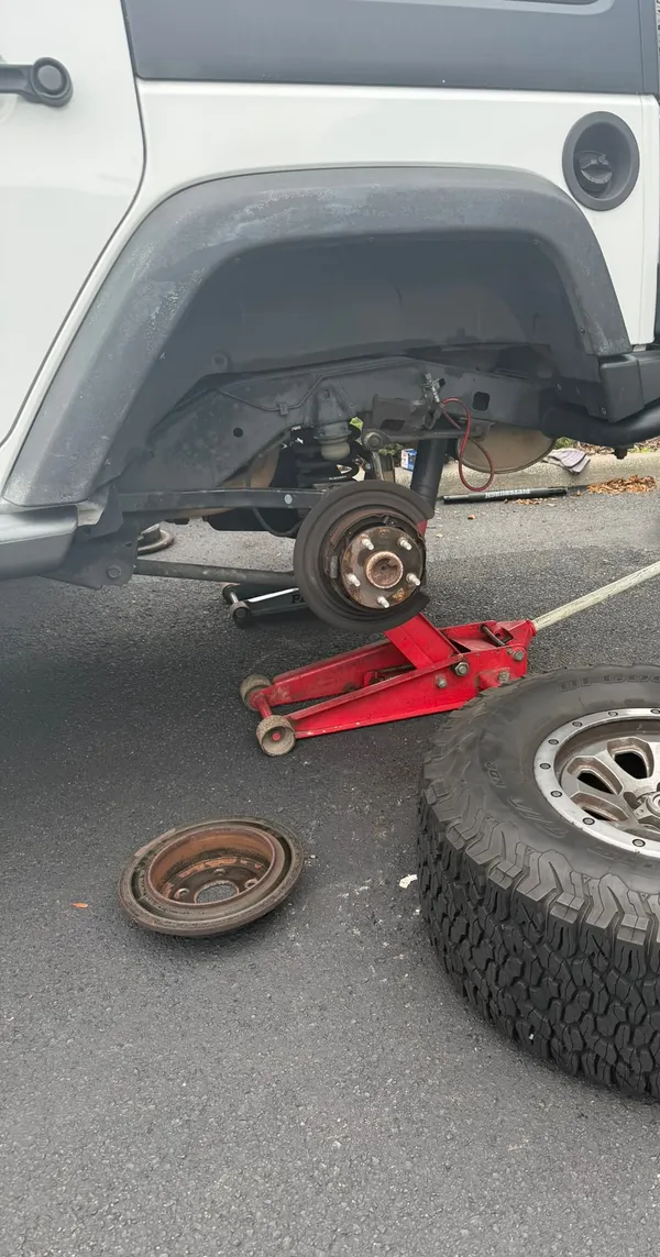 Rear wheel removed from a white Jeep Wrangler on a red floor jack, with the worn rotor still on the hub and the old rotor and the wheel lying on the asphalt