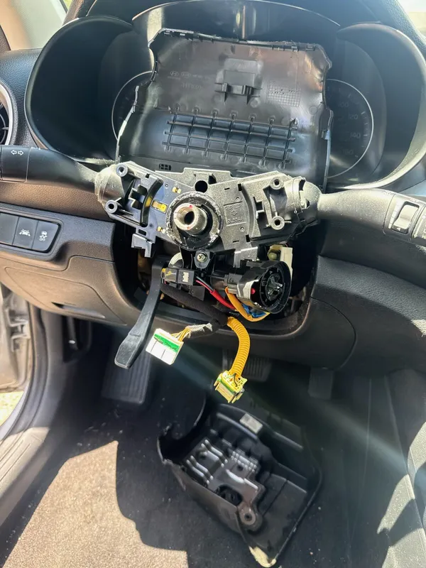 Kia steering column with the wheel and shrouds off and the cluster hood lifted, the damaged ignition below the switch housing and the lower shroud on the floor mat