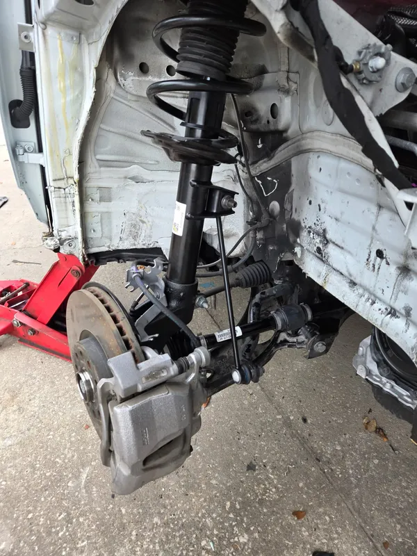 Front corner of a white car with the wheel off, showing the strut, brake rotor, caliper and tie rod, with the body panels already removed and a red jack under the sill