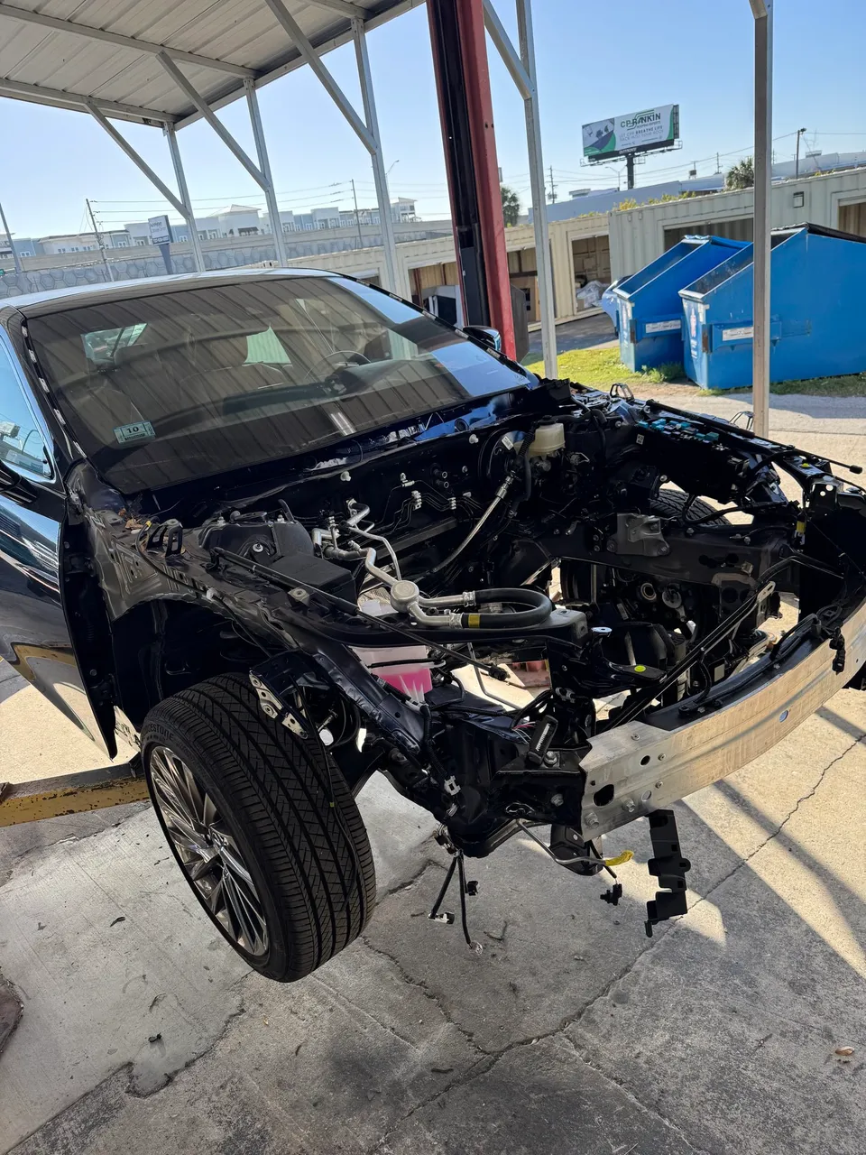 Black Lexus with its front clip and engine removed, seen from the front corner, on stands at a Sarasota collision center