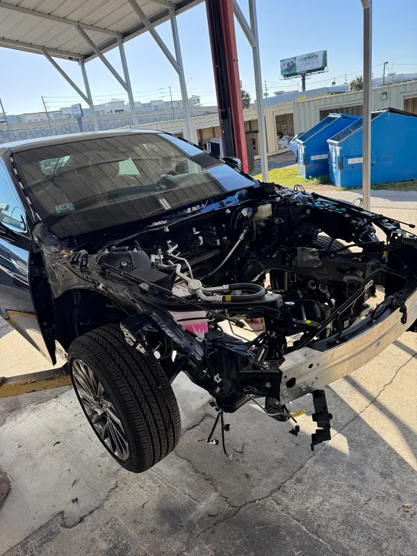 Black Lexus with its front clip and engine removed, seen from the front corner, on stands at a Sarasota collision center