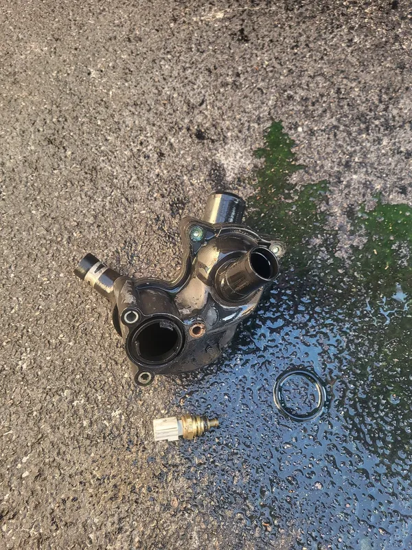 The removed thermostat housing on wet asphalt beside a coolant puddle, with its temperature sensor and seal laid out next to it