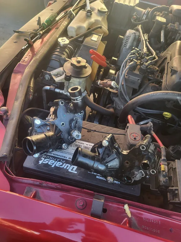 The new thermostat housing and the old one side by side on top of the battery, the old one dark with coolant and grime