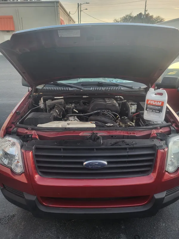 Red Ford Explorer with the hood up in a parking lot outside a commercial building, a jug of antifreeze resting on the radiator support