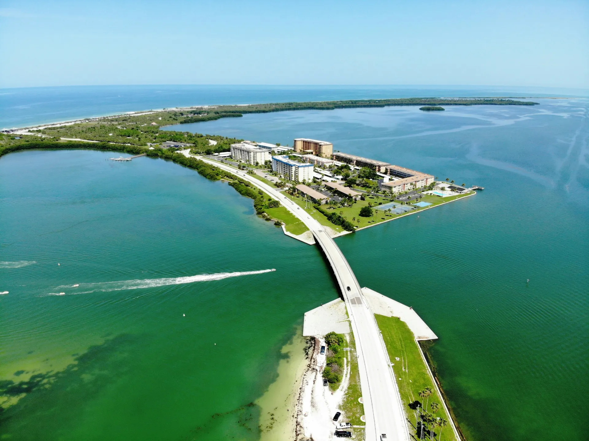 Aerial view of the Dunedin Causeway crossing shallow turquoise water toward waterfront condominium buildings and the barrier island beyond