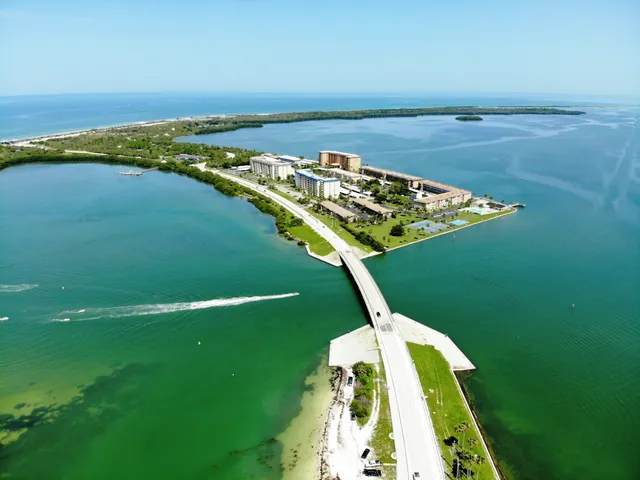 Aerial view of the Dunedin Causeway crossing shallow turquoise water toward waterfront condominium buildings and the barrier island beyond