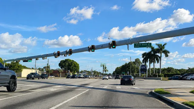 Traffic crossing the Park Boulevard North intersection in Pinellas Park, signal heads hanging from the mast arm overhead, palms and the SPC campus sign on the corner
