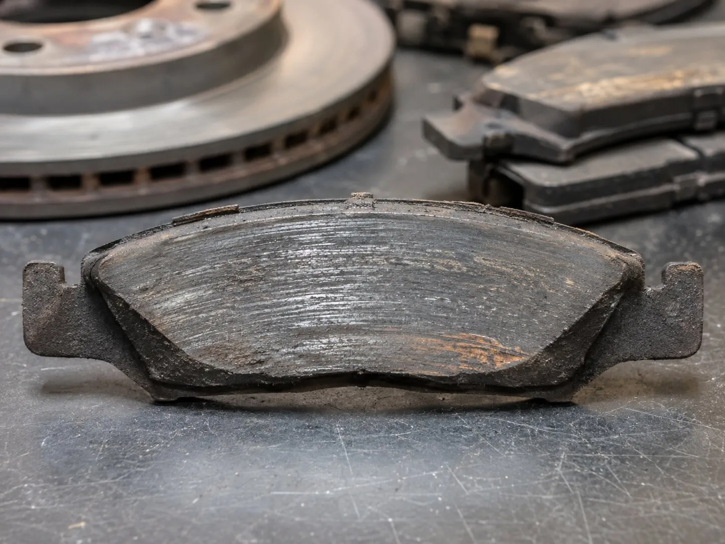 A worn brake pad standing on a bench, its friction material ground thin and scored, with a used vented rotor and a stack of other spent pads behind it