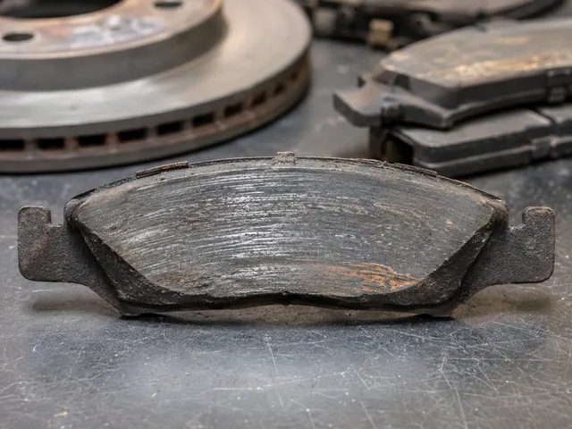A worn brake pad standing on a bench, its friction material ground thin and scored, with a used vented rotor and a stack of other spent pads behind it